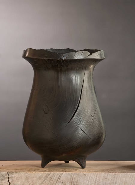 Charred wooden art vessel with three feet, cracked rim, and rich blackened surface texture.